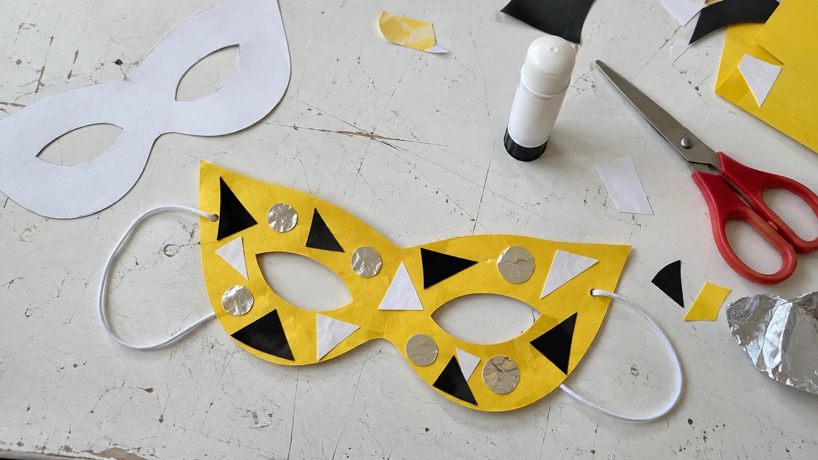 Zelfgemaakt geel papieren carnavalsmasker met zwart-witte versiering, schaar en papierrestjes